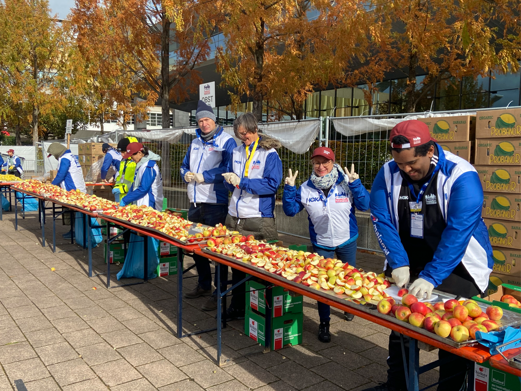 Ehrenamtliche in blauen und weißen Jacken bereiten im Freien an langen Tischen geschnittene Äpfel für einen Marathon vor.