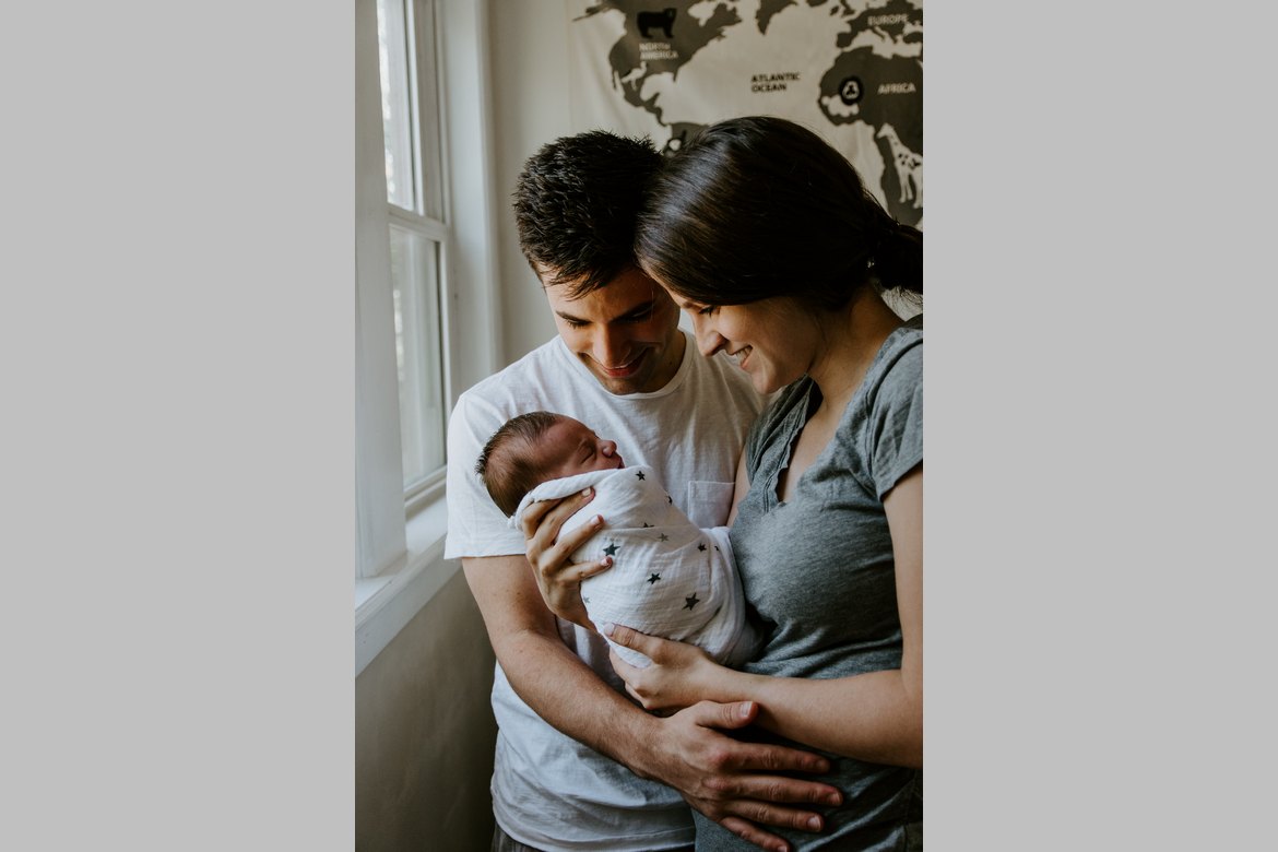 Junge Fau und junger Mann schauen liebevoll auf Neugeborenes, das die junge Frau im Arm hat.