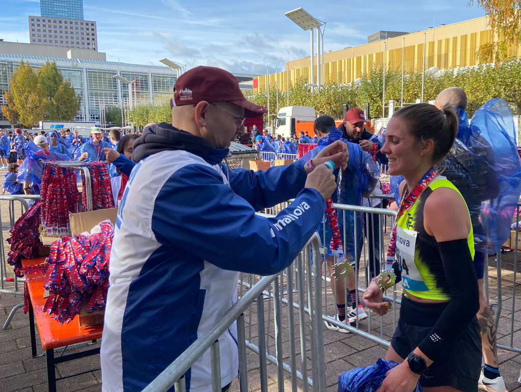 Ein Helfer in blau-weißer Jacke hängt einer lächelnden Läuferin in Sportkleidung nach einem Rennen im Freien eine Medaille um.