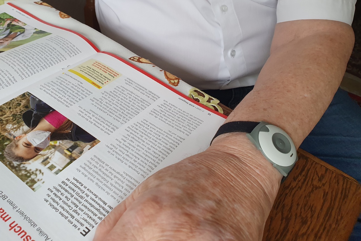 Arm mit Hausnotrufknopf liegt auf einer Zeitschrift auf dem Tisch 