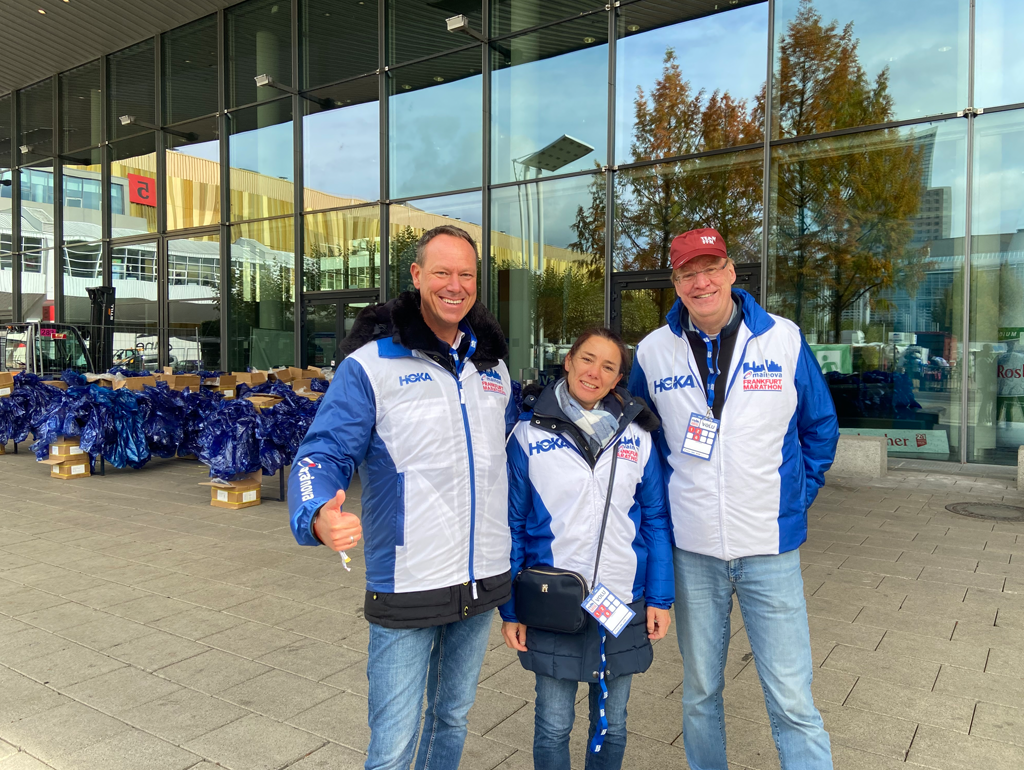     Anfrage erfolgreich  Drei lächelnde Freiwillige in blauen und weißen Marathon-Jacken posieren gemeinsam im Freien vor einer modernen Glasfassade.