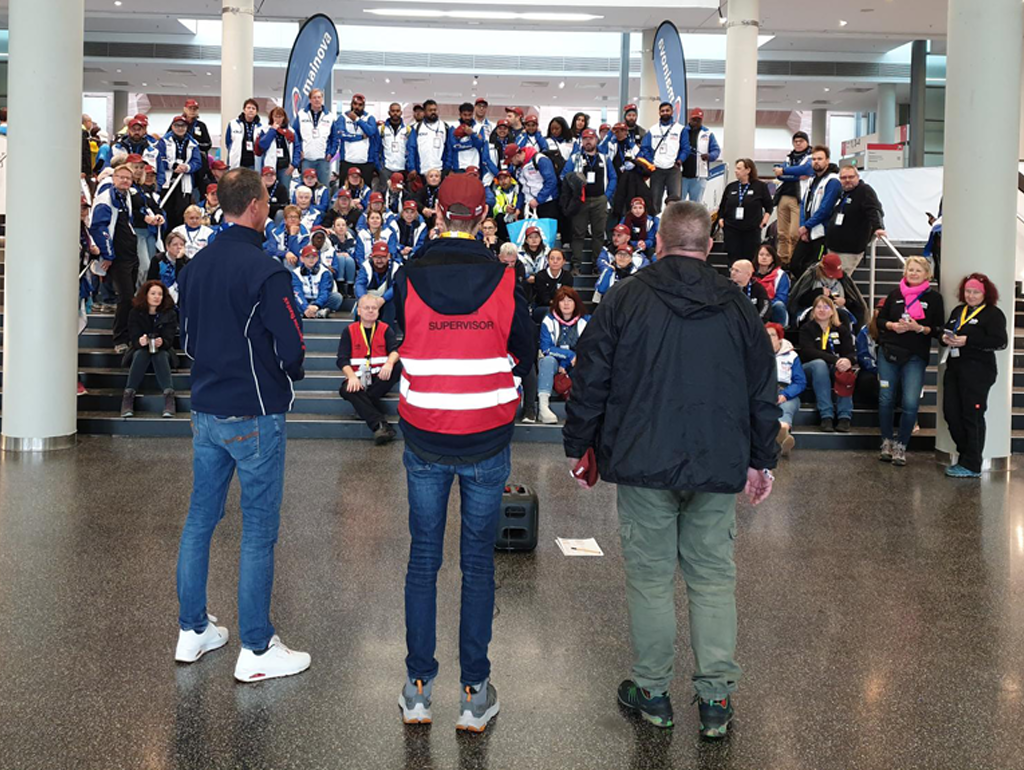 Drei Personen stehen im Vordergrund mit dem Rücken zur Kamera in einer großen Halle. Im Hintergrund sitzt eine große Gruppe von Menschen in blauen und weißen Teamjacken auf einer breiten Treppe.