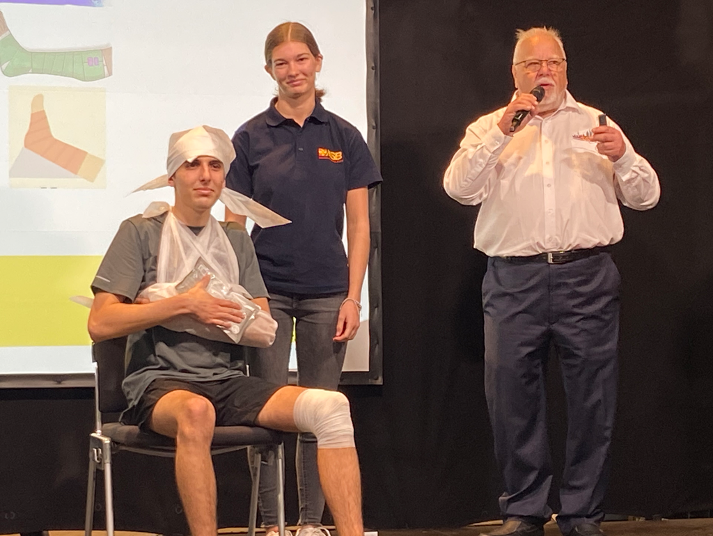 Drei Personen bei einer Erste-Hilfe-Demonstration: Ein Mann mittleren Alters spricht ins Mikrofon, während eine junge Frau hinter einem jungen Mann steht, der mit Verbänden und einer Notfalldecke auf einem Stuhl sitzt.