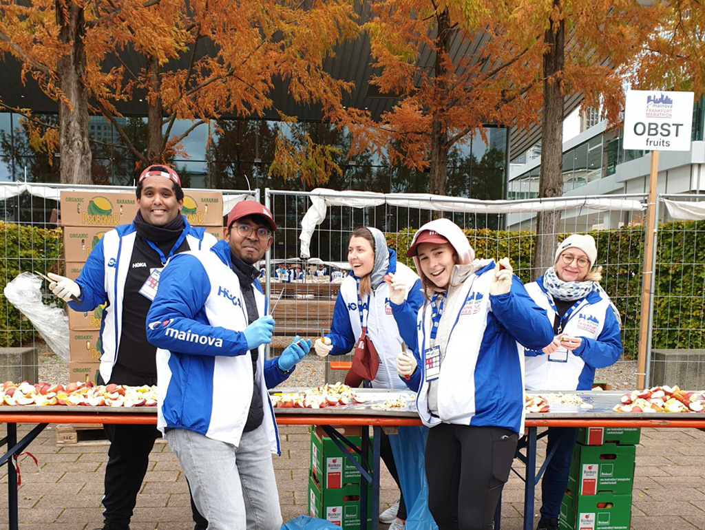Fünf lächelnde Helfer in blauen und weißen Jacken stehen hinter einem Tisch mit geschnittenen Äpfeln für den Marathon.