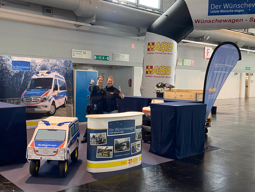 Zwei lächelnde Frauen stehen an einem Informationsstand des "Wünschewagen - Letzte Wünsche wagen" in einer großen Halle. Im Hintergrund ist ein großes aufblasbares Banner mit dem Logo des ASB (Arbeiter-Samariter-Bund) zu sehen.
