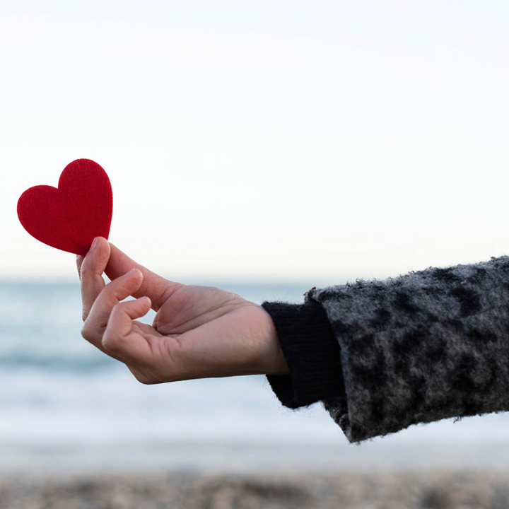 Eine Hand hält ein rotes Papierherz vor einem unscharfen Hintergrund mit Meer und Himmel.